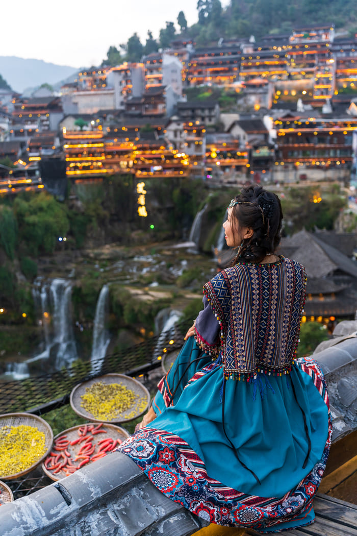 Mujer con vestimenta tradicional contemplando el pueblo antiguo de Furong iluminado y sus cascadas al atardecer en China.